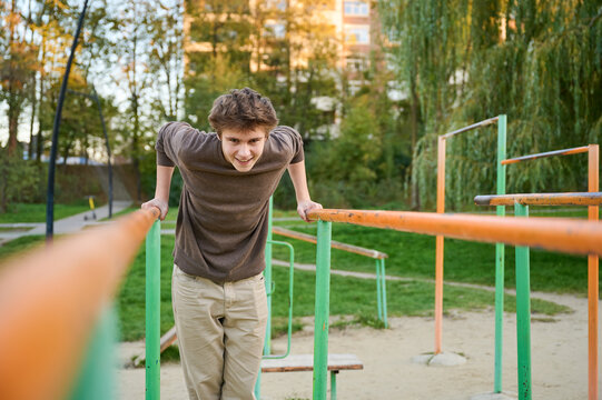 White athletic man performing parallel dips with controlled posture, outdoor calisthenics park setting with orangegreen equipment, intense focused gaze, apartment blocks and trees in background,