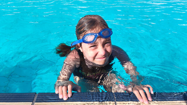A girl in swimming goggles emerges from the water near the edge of a pool. The idea is for summer vacations, active childhood, swimming lessons, and a healthy lifestyle.