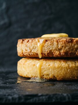 Side Profile View of Two Stacked Cassava Bammy Rounds with Melting Butter Pat Dripping Down, Dark Slate Background

