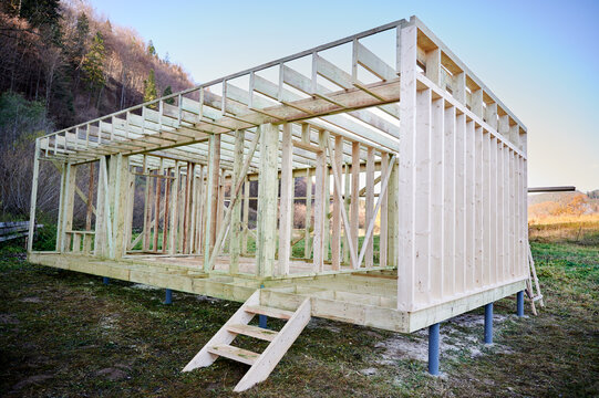 Wooden frame house under construction on pile foundation in open landscape, unfinished timber structure symbolizing residential development, prefab building, investment and growth