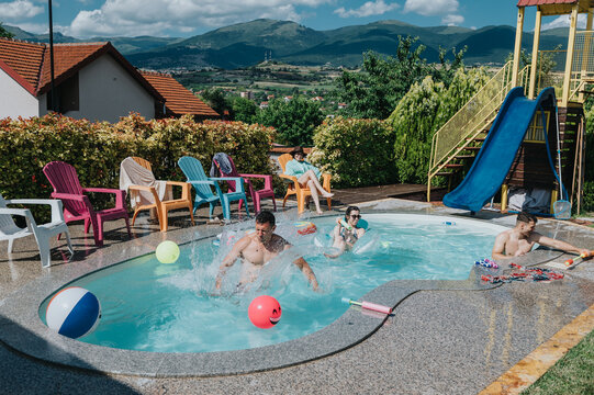 A group of friends and family play and relax in a small backyard swimming pool. Sunlit summer fun by a pool with chairs, floaters and a water slide nearby.