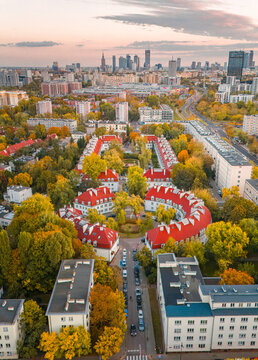 Aerial view of residential buildings with bright red roofs surrounded by autumn trees and the modern city skyline under a soft sunset sky in Poland.