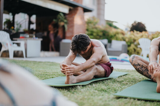 A shirtless man reaches forward to stretch his hamstrings on a yoga mat. Other participants practice stretches outdoors in a relaxed backyard fitness session.