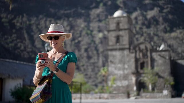 Happy senior woman smiles while checking her selfies on her phone at San Javier Mission near Loreto at sunrise in Baja California Sur