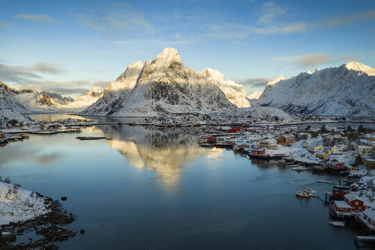Aerial view of snow-capped mountains and a traditional coastal village with red houses reflected in the calm blue water of a fjord Norway.