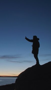 Silhouette of a man standing on a rocky shore and taking a selfie with a smartphone at twilight. Dark figure against a deep blue evening sky and calm water creates a dramatic outdoor travel scene with