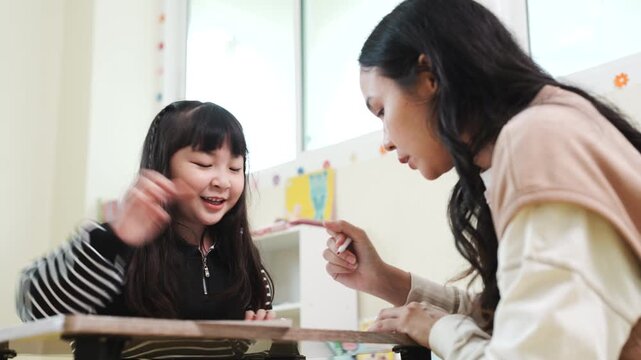 Happy Asian little girl sharing a marker with her teacher in classroom, early childhood education and fun learning concept.