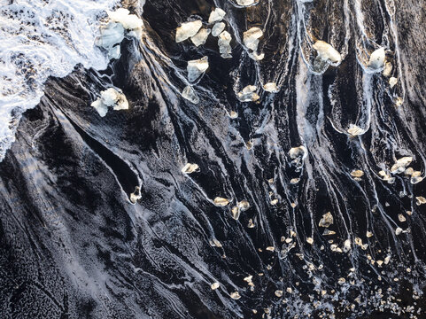 Aerial view of glacial ice chunks scattered across the black sand of Diamond Beach with white ocean foam washing ashore in Iceland.