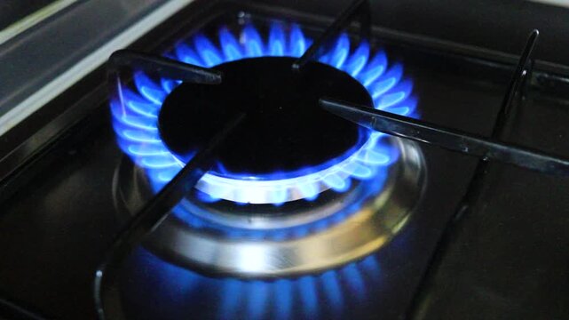 A slow-motion shot of a blue flame igniting on a gas burner on a kitchen stove. A close-up of a household gas stove in a dark room. Energy consumption during cooking, gas consumption, utility bills