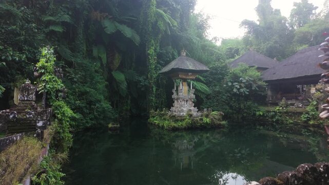 Balinese Hindu pelinggih shrine sits on the water with a backdrop of jungle and crystal clear water. Slow motion camera track forwards. Location: Pura Gunung Kawi Sebatu Water Temple, Ubud, Bali