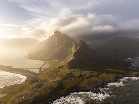 Aerial view of a dramatic mountain peak shrouded in clouds with a winding coastal road and waves crashing against the rocky shore during golden hour Iceland.