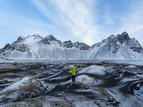 Aerial view of a person in a yellow jacket standing on snow-dusted black sand dunes with arms raised towards the majestic Vestrahorn mountain range in Iceland.