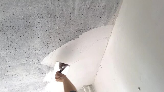 hand of male worker applying white putty to grey textured ceiling surface with metal trowel during interior renovation, copy space. Home improvement, construction work, wall finishing, repair project