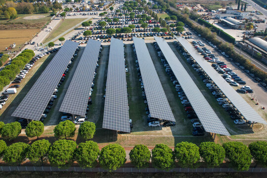 Aerial view of long rows of solar panels covering parked cars at the Fiera di Rimini exhibition center surrounded by green trees in Rimini, Emilia-Romagna, Italy.