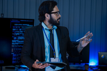 South Asian male developer concentrates silhouetted against bright monitor screens showing...
