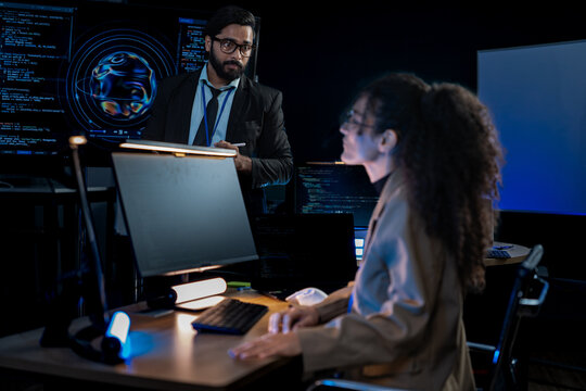 South Asian male and Black female software developers pair-program at dual monitors displaying network security algorithms at night.