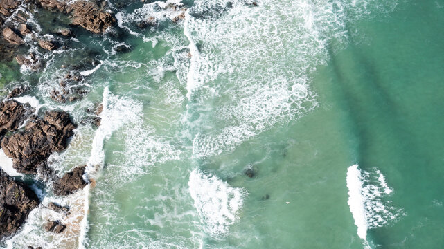 Aerial view of turquoise waves crashing against dark coastal rocks and sandy patches at Trevone Bay Padstow, Cornwall, United Kingdom.