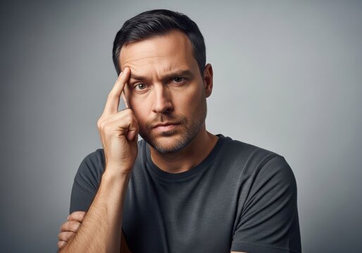 Portrait of a thoughtful man with a serious expression touching his forehead, looking directly at the camera against a neutral grey background