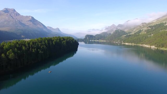 A serene panoramic view of a tranquil glacial lake surrounded by lush green forests and towering rocky mountain peaks under a clear blue summer sky