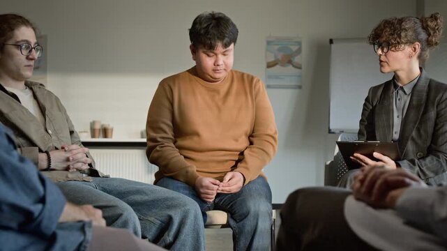 Caucasian female psychotherapist taking notes with Asian young man sharing feelings in support group. Clip for mental health awareness and therapy service commercials