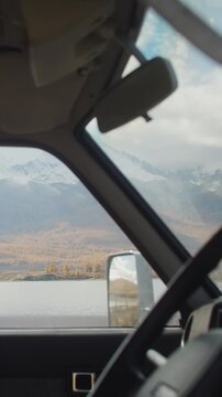Timelapse view from inside a car looking through the window at a calm lake, autumn forest, and snowy mountain valley under a cloudy sky. Vehicle interior frames the remote natural landscape, creating
