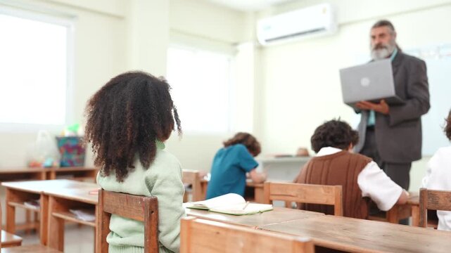 Girl raising hand in diverse elementary classroom to ask teacher a question. Student sitting at wooden desk participating in learning lesson.