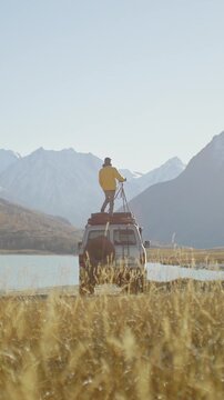 Photographer in a yellow jacket standing on the roof of an off road SUV with a camera on a tripod by a mountain lake. Golden grass, bright sunlight, and snowy alpine peaks create a dramatic outdoor