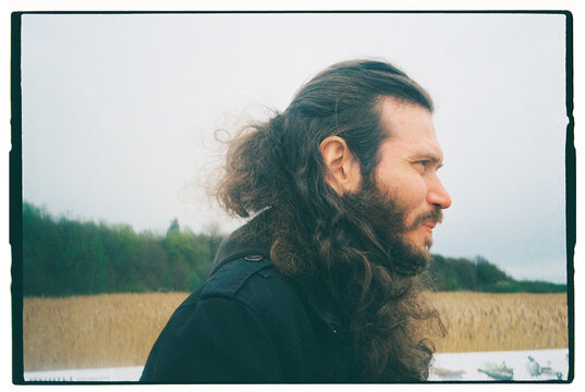 Man with long hair stands by tall grass on cloudy day