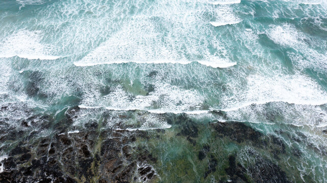 Aerial view of turquoise ocean waves with white foam crashing against dark rocky cliffs at Constantine Bay in Padstow, Cornwall, United Kingdom.