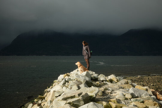 Person stands with dog on rocks near water and mountains on cloudy day