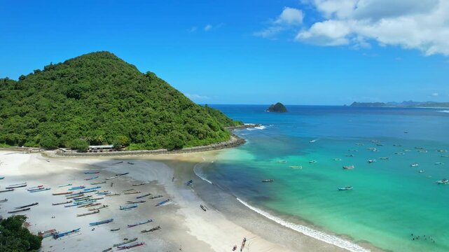 Aerial drone footage of Selong Belanak beach, Lombok island, Indonesia, white sand beach bay, crystal turquoise blue waters, conic green hill, beach restaurants and bars, surfers, many fishing boats