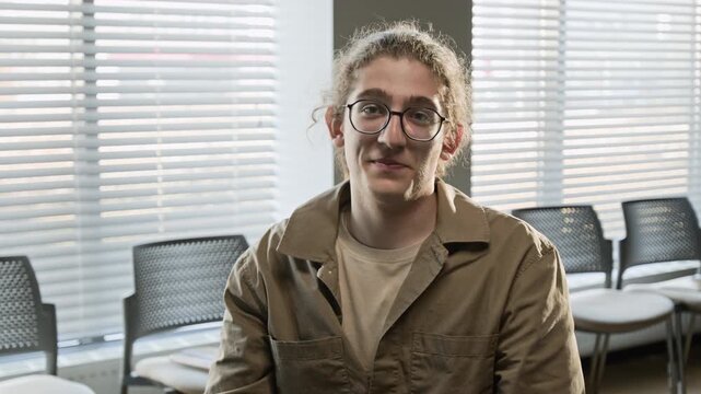 Chest-up portrait of Caucasian male teenager with glasses looking at camera and smiling in group therapy room. Clip for youth mental health campaigns and counseling services