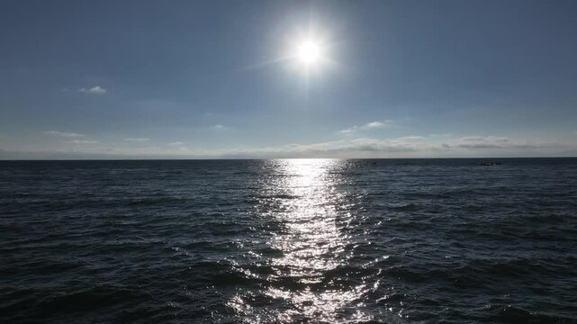 The sun shines brightly on the deep blue sea, creating sparkling reflections on the gentle waves as a low altitude drone takes forward flight