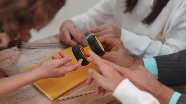 Close up of teacher and student hands pointing at robot wheel. Learning STEM science and engineering project together on wooden desk.