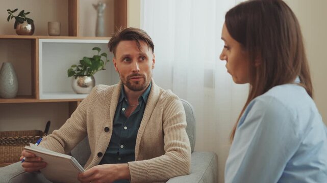 Male psychologist taking notes while listening to female client during therapy session in office with calm supportive atmosphere