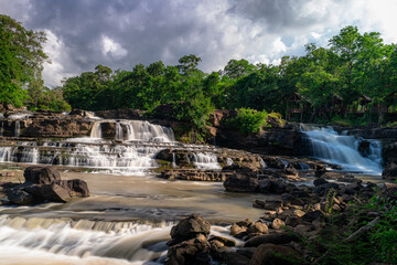 Long exposure photography of Tad Hang Waterfall in Laos. Multiple cascades of silky white water flow over wide rocky tiers into the river below, surrounded by dense tropical rainforest and ancient tre © jr