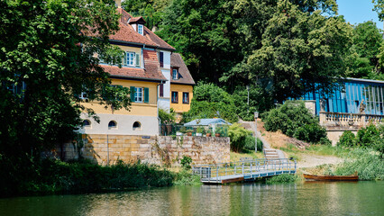 Fototapeta premium Scenic riverfront view with a historic yellow house, a small pier, and a wooden boat on a green forest bank in Bamberg, Germany.