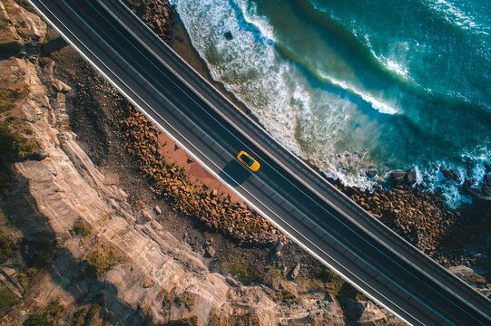 Small yellow car driving along the scenic coastal highway bridge captured from a high aerial perspective during a sunny day