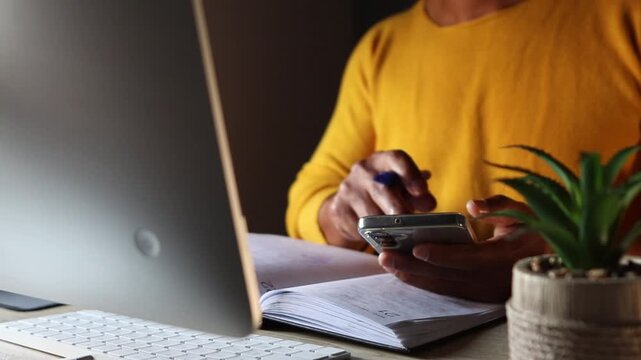 Person working at desk using smartphone and writing notes