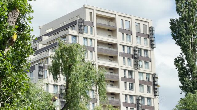 A large modern residential building rises proudly into the sky behind lush green trees. The bright daylight creates a peaceful, comfortable feeling of contemporary urban living.