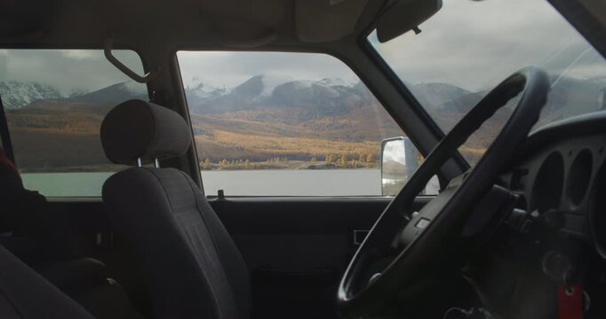 Timelapse view from inside a car looking through the window at a calm lake, autumn forest, and snowy mountain valley under a cloudy sky. Vehicle interior frames the remote natural landscape, creating