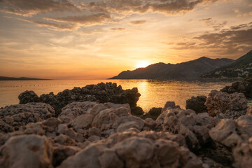 Fototapeta premium Golden sunset over the Adriatic Sea in Brela, Croatia. Rocky coast in the foreground and mountains on the horizon during golden hour on the Makarska Riviera.