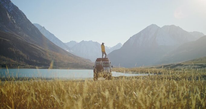 Traveler in a yellow jacket standing on the roof of an off road SUV by a mountain lake and looking across a wide alpine valley toward snowy peaks. Golden grass, bright sunlight, and remote wilderness