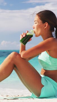 Fitness woman drinking a healthy green smoothie or cold pressed juice for a detox weight loss cleanse. Happy sporty asian girl on beach taking a snack break vegan beverage. Vertical video
