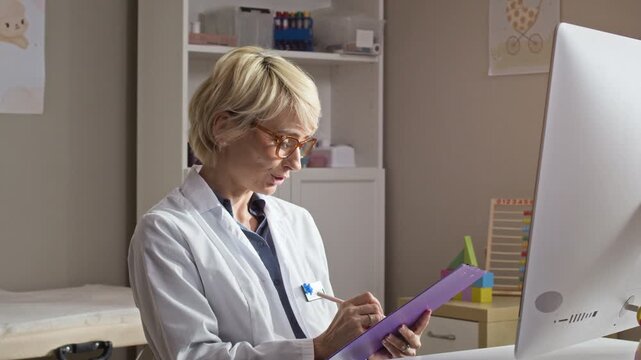 Waist up of middle aged Caucasian female pediatrician wearing glasses and lab coat talking during online consultation and writing notes on clipboard. Ideal for telehealth and digital medicine ads