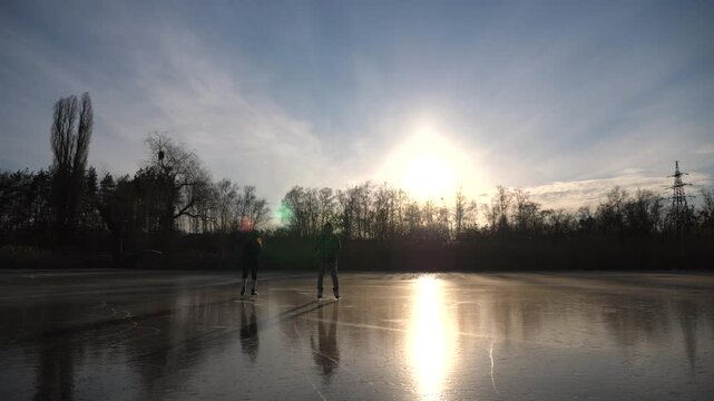 Two young people in figure skates sliding on ice during sunset. Man and woman skating on frozen river. Shining sun is reflecting on surface of ice. Couple have active leisure at winter. Slow motion