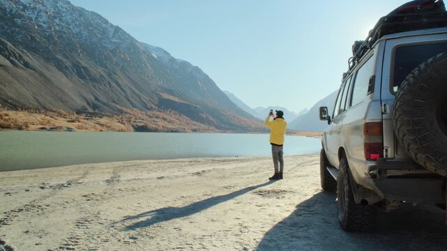 Man in a yellow jacket photographing a mountain lake with a smartphone while standing near an off road vehicle on a sandy lakeshore. Wide alpine landscape, bright sun, and remote wilderness atmosphere