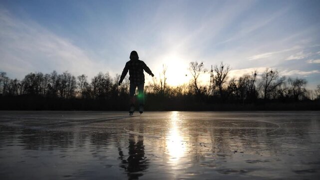 Young man shod in figure skates sliding on ice surface during beautiful sunset. Sportsman skating on frozen river at freeze day. Male skater having active leisure at lake in winter cold weather