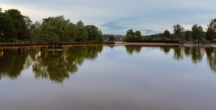 A wide panoramic shot of a calm pond reflecting lush green trees and a wooden bridge in the famous Zdrojowy Park in Kudowa Zdroj