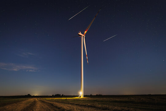 View of a tall wind turbine standing in a rural field under a starry night sky with light trails in Masovian Voivodeship, Poland.
